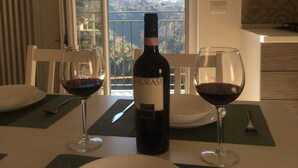 Une table à manger avec une bouteille de vin, deux verres et des assiettes préparées pour un repas. Une fenêtre offre une vue sur un paysage montagneux.