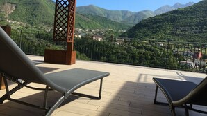 Une terrasse avec deux fauteuils de jardin, une pergola en bois et une vue sur un paysage montagneux avec des bâtiments dans la vallée.