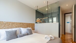 Quarto de hotel moderno com uma cama grande, cabeceira de madeira, divisória de vidro e piso de madeira.