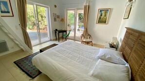 Ein Schlafzimmer mit einem großen Bett, einem Schreibtisch und Stühlen. Es gibt zwei Fenster mit Vorhängen, eine Tür und eine Wandlampe.