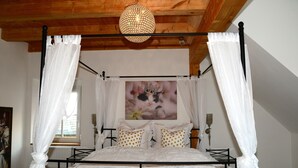 Una cama con dosel y cortinas, con una colcha blanca y almohadas con lunares dorados, un cuadro en la pared y un techo de madera.