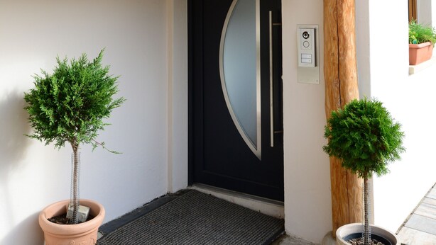Una escalera de madera que conduce a una puerta negra con diseño moderno, dos plantas en macetas y una barandilla metálica.