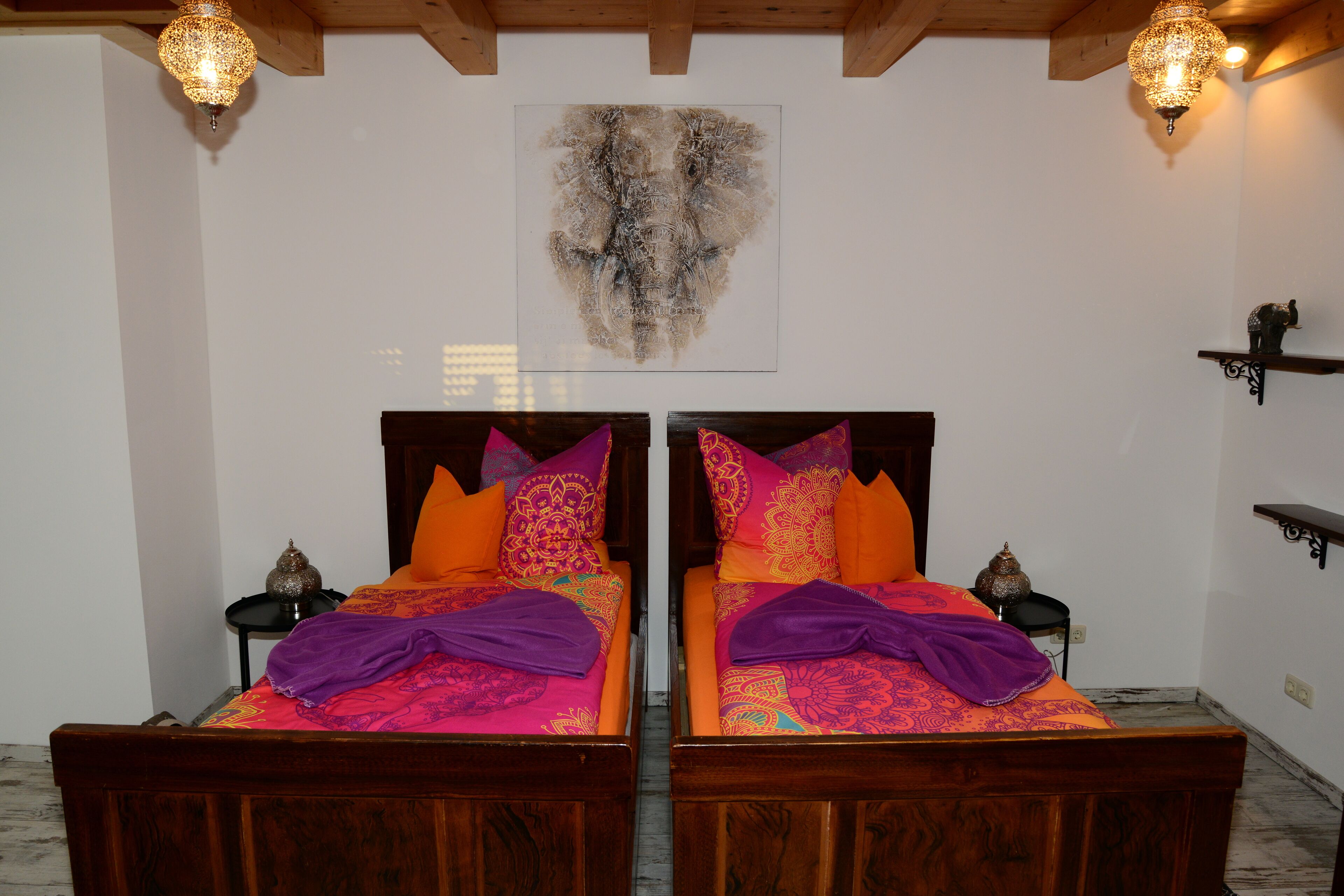 Una habitación con dos camas de madera, cada una con ropa de cama colorida y mantas púrpuras. Hay luces fijadas a la pared y un cuadro abstracto grande sobre las camas.