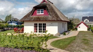 A thatched-roof house with a white exterior and red accents, surrounded by a well-maintained garden and a paved driveway.