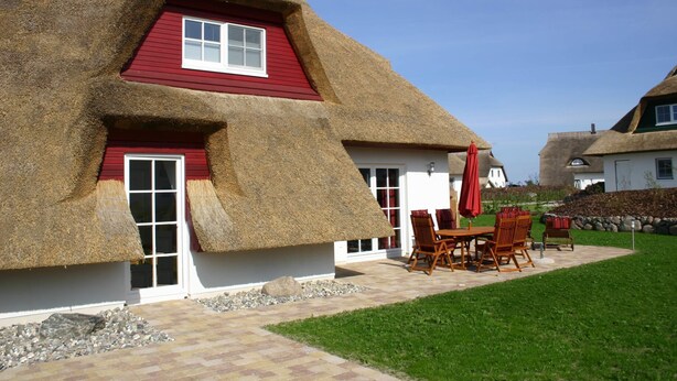 A house with a thatched roof and red trim, a patio with outdoor furniture, and a well-maintained lawn.
