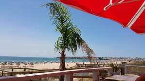 En vy över en strand med en palm, strandparasoller och en stadsbild i bakgrunden.