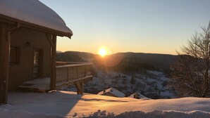 Une cabane en bois, dont le toit est recouvert de neige, avec un balcon donnant sur un paysage enneigé au coucher du soleil.