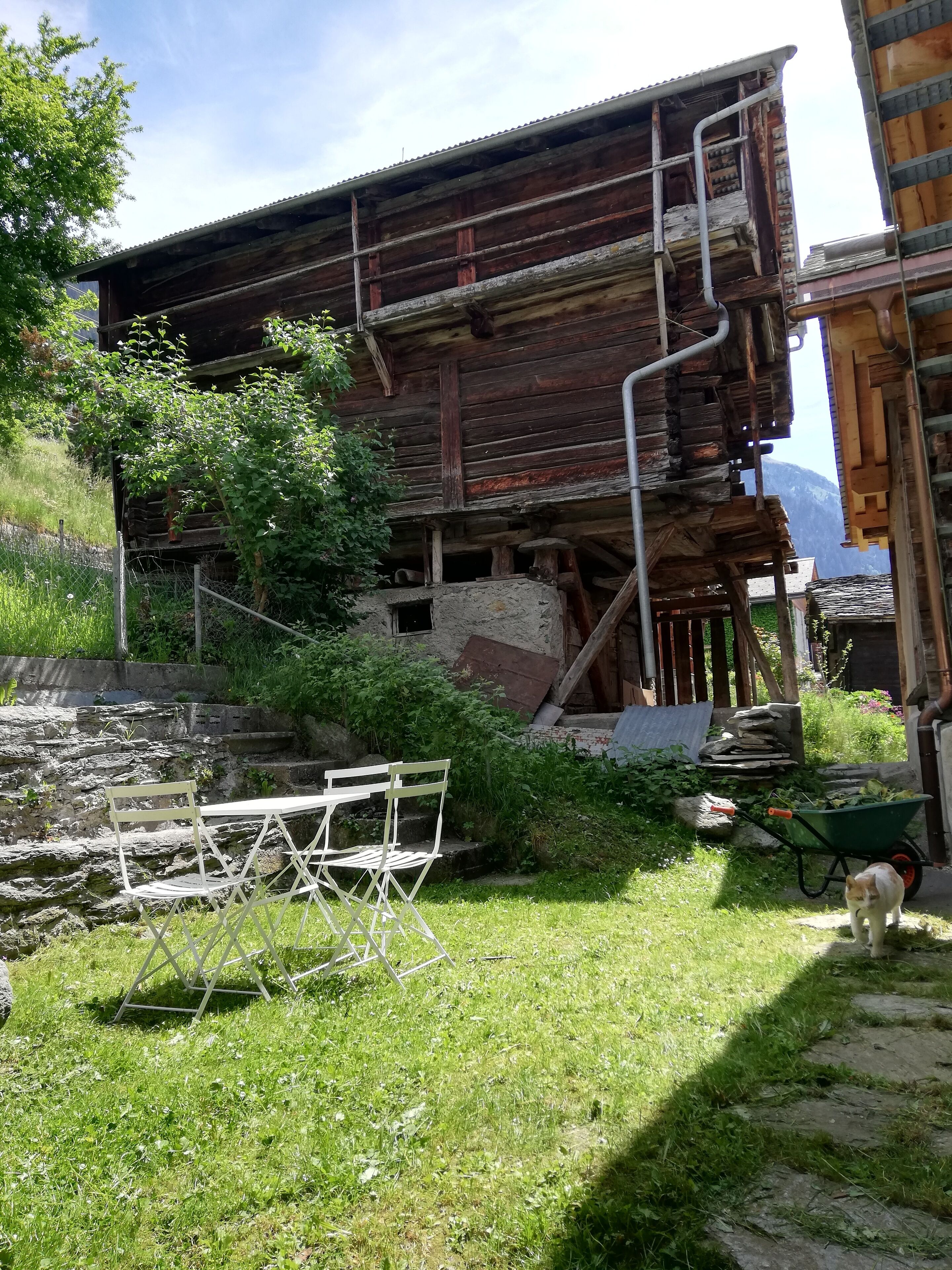 Ein rustikales Holzgebäude mit einer Metalltreppe, einer Steinmauer und einer Rasenfläche mit Klappstuhl und Schubkarre.