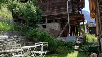 Ein rustikales Holzgebäude mit einer Metalltreppe, einer Steinmauer und einer Rasenfläche mit Klappstuhl und Schubkarre.