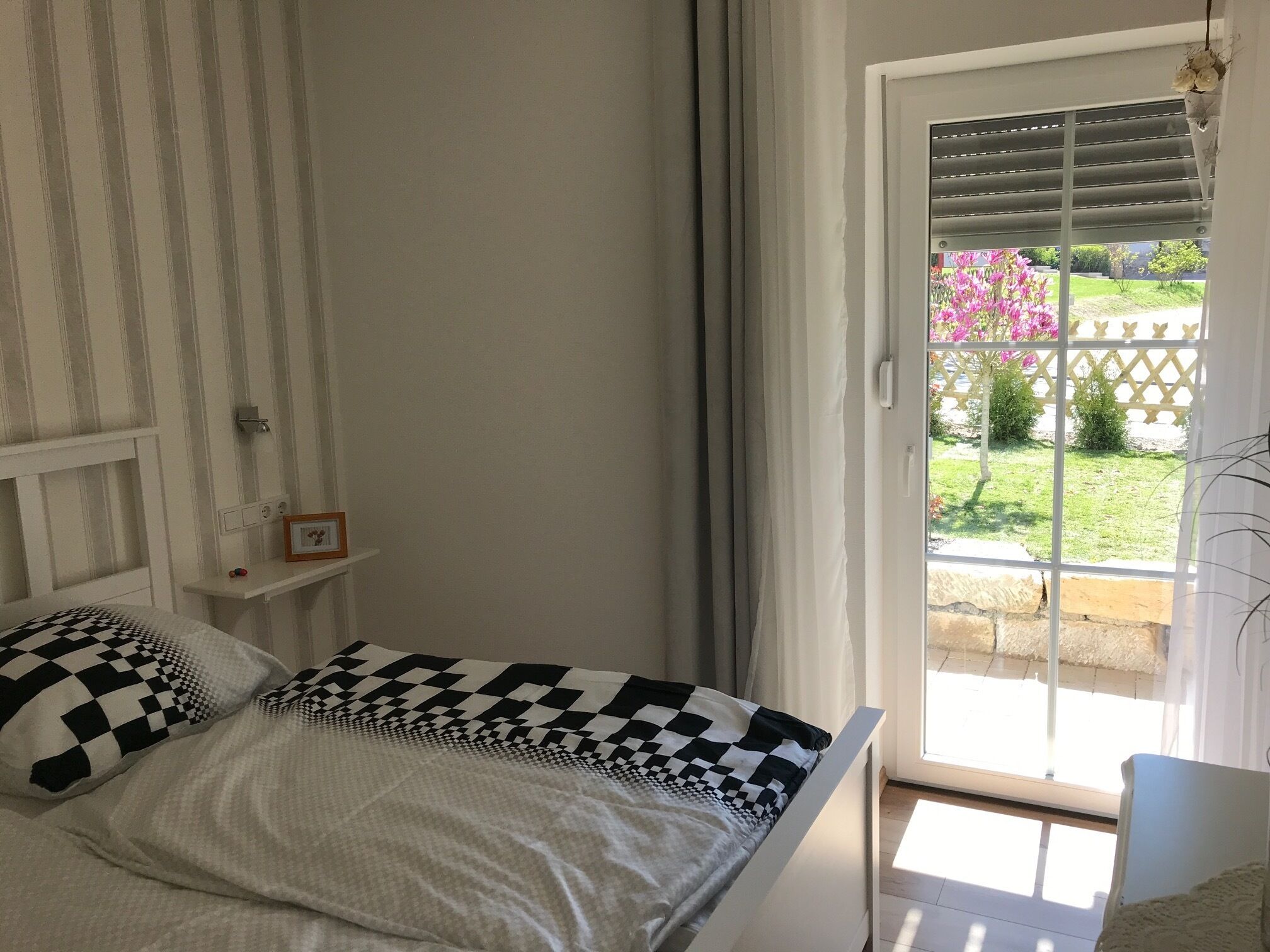A bedroom with a bed, a bedside table, a window with curtains, and a view of a garden.
