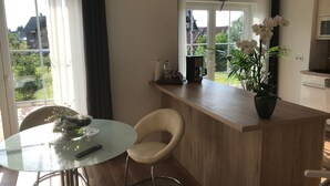 Eine moderne Küche mit einer Holzarbeitsplatte, einem runden Glastisch und einem Stuhl. Auf der Arbeitsplatte steht ein Blumentopf und es gibt ein Fenster mit Blick ins Freie.