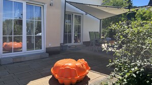 Eine Terrasse mit einer großen, orangefarbenen Schildkröten-Skulptur, einem überdachten Sitzbereich und einem Haus mit großen Fenstern und einem Ziegeldach.