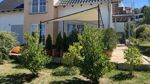 Ein zweigeschossiges Haus mit einem Ziegeldach, einem Balkon mit einer Pergola und einem Garten mit Topfpflanzen.