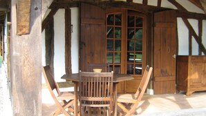 A wooden table and chairs set up under a covered area with a large window and wooden doors. - In the heart of the Landes forest, traditional half-timbered gîte, 4pers. (Argelouse)
