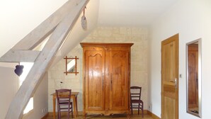 Ein Schlafzimmer mit schrägen Wänden, einem Holzschrank, zwei Stühlen, einem Bett mit gestreifter Bettdecke und einem kleinen Tisch mit einer Vase.
