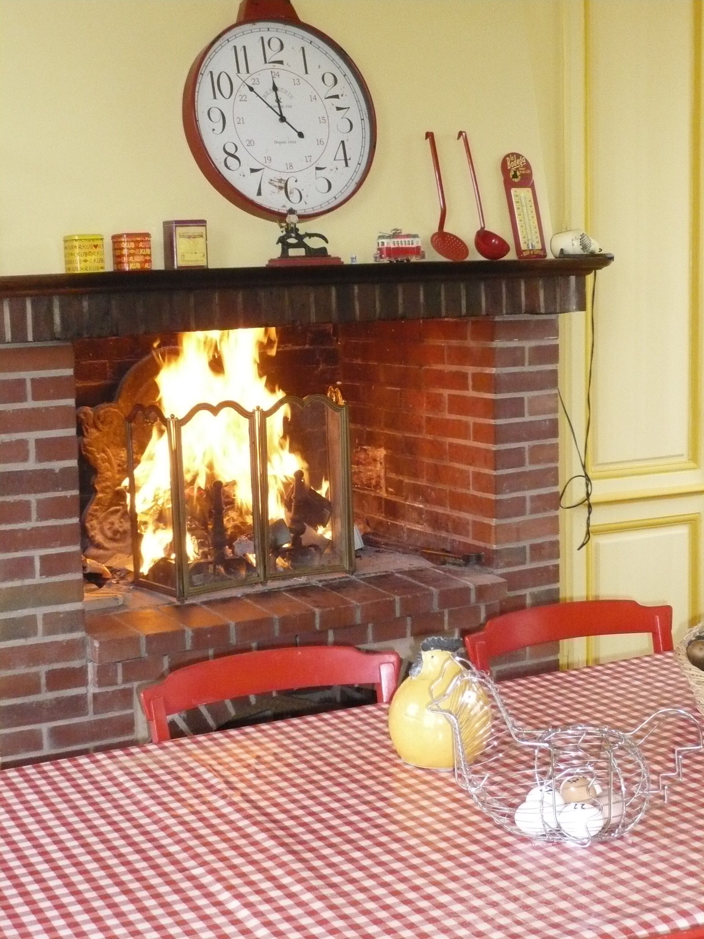 Une cheminée surmontée d’une horloge, une table recouverte d’une nappe à carreaux et des chaises disposées autour de la table.