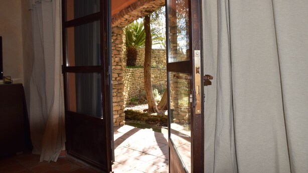 An open door leading to an outdoor area with a stone wall, a palm tree, and a tiled floor. - An independent apartment in a pretty property in the countryside ... (CORNILLON CONFOUX)