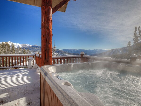 Relax in a hot tub on a deck surrounded by snow-capped mountains and evergreen trees.