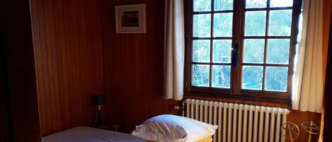 Ein Zimmer mit zwei Betten, einem Heizkörper und einem fensterbelaubten Fenster.
