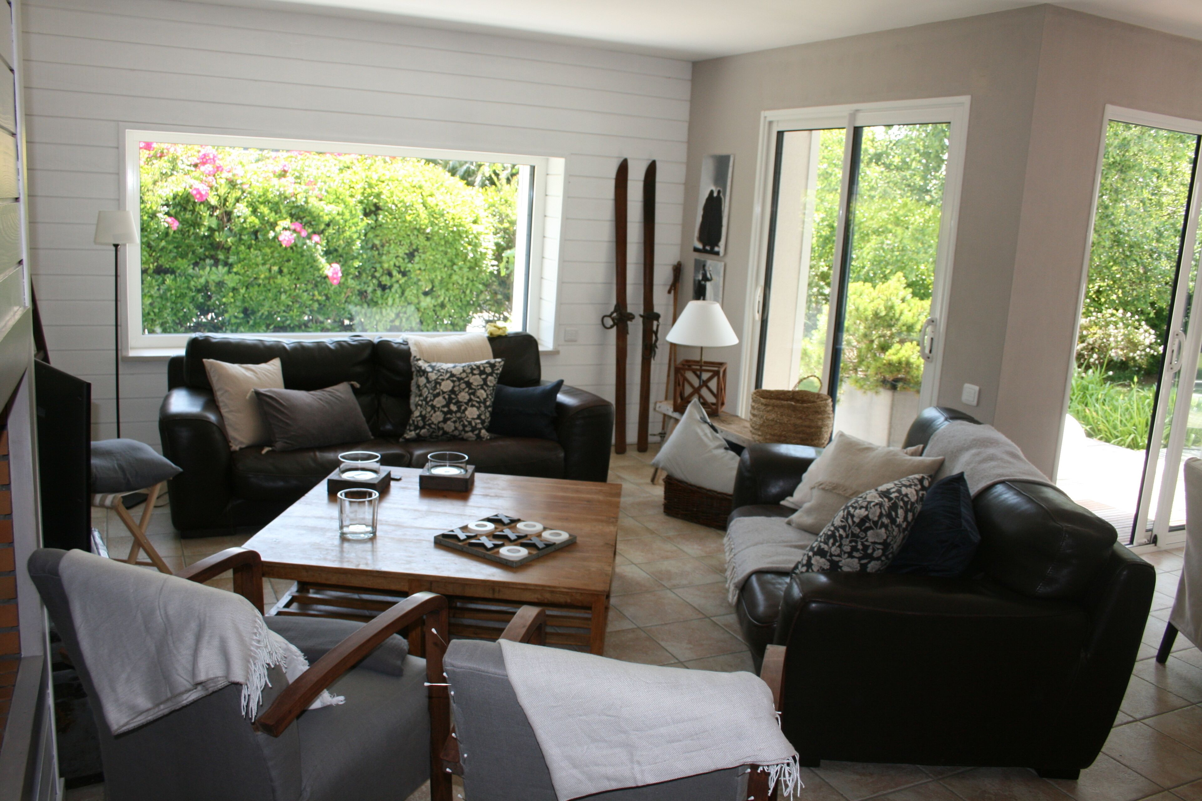 A living room with a sofa, a coffee table, and a dining table. There are two windows with a view of greenery outside.