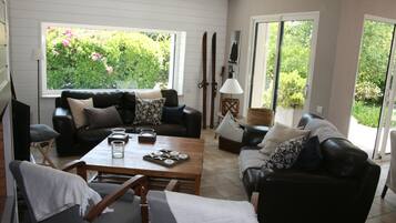 A living room with a sofa, a coffee table, and a dining table. There are two windows with a view of greenery outside.
