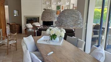 A modern dining area with a wooden table, white chairs, and a vase of white flowers. There is a fireplace, wicker lamps, and a view of greenery outside.