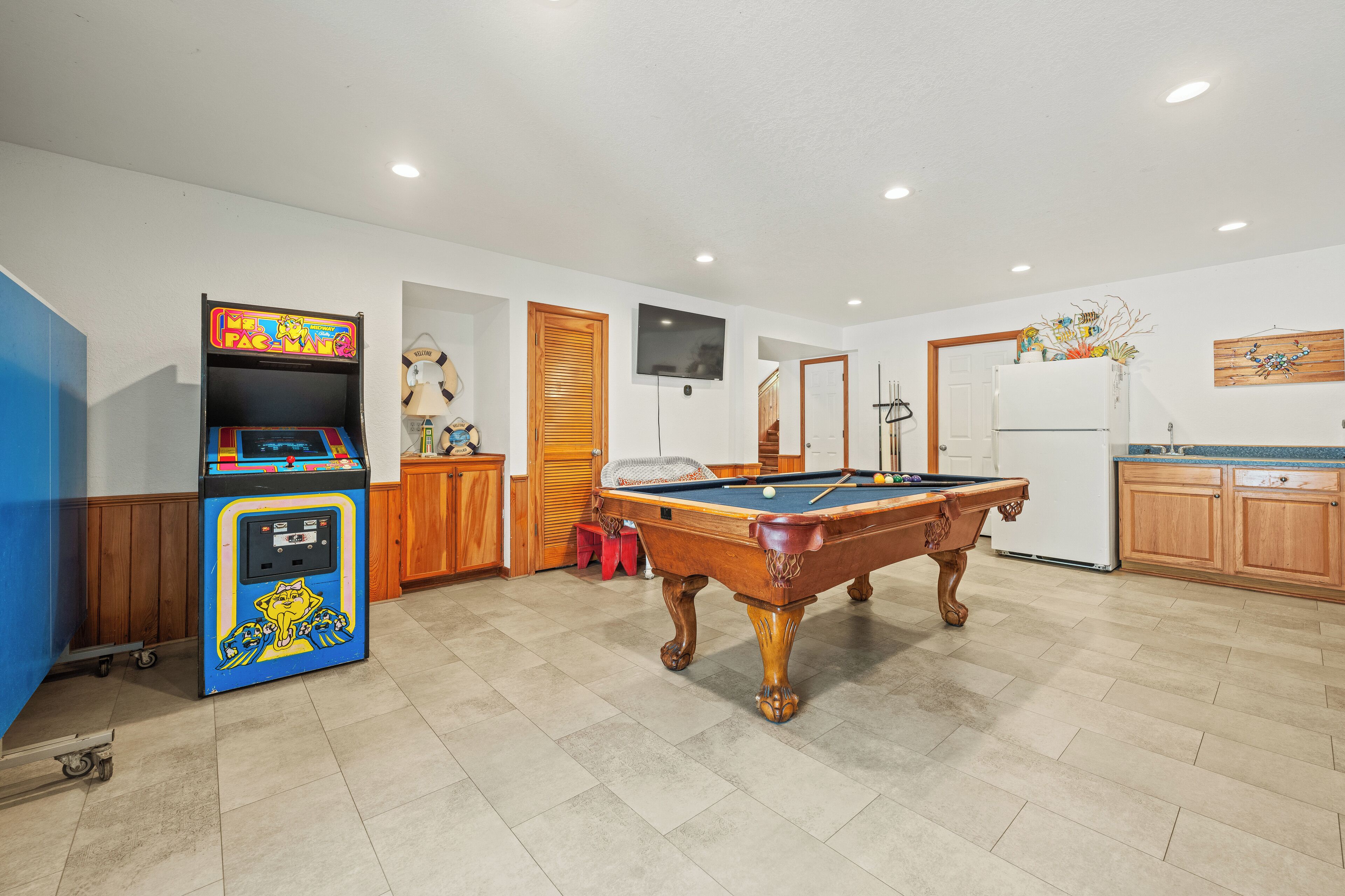 A room with a pool table, a video game machine, a television, and a refrigerator.