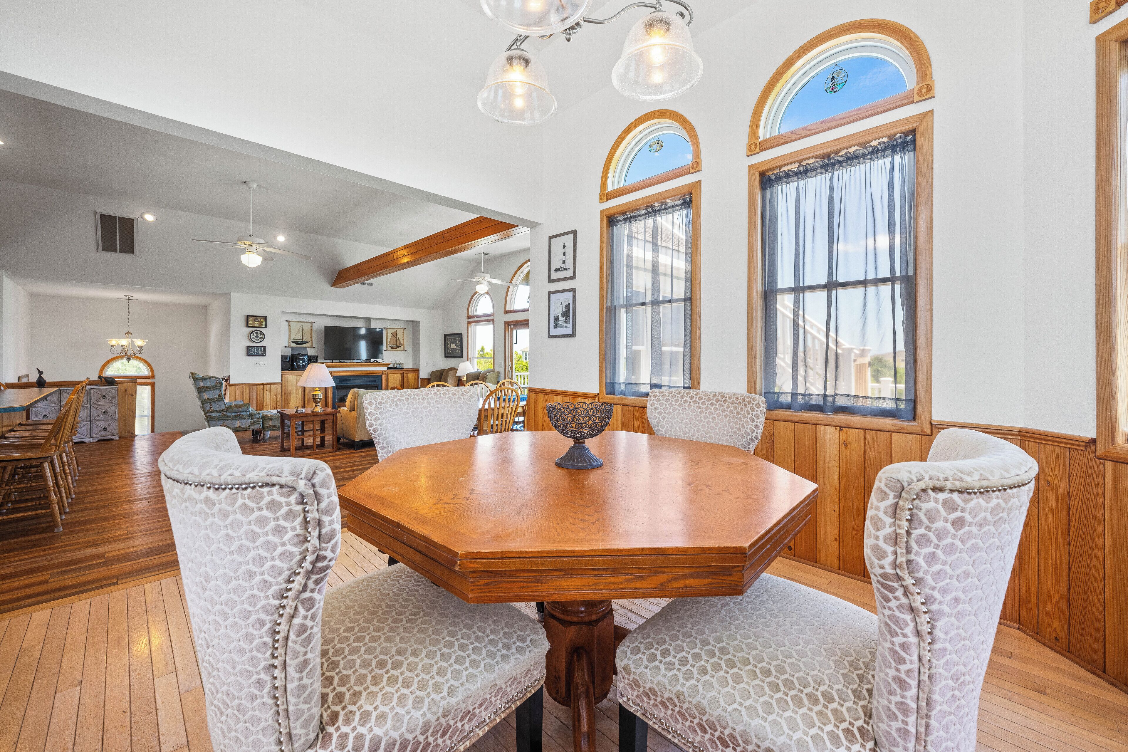 A dining area with a wooden table and patterned chairs, large windows with curtains, and a kitchen in the background.