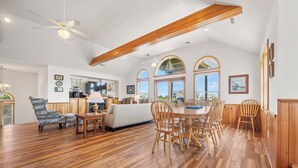 A spacious living room with a wooden floor, a dining area with wooden chairs, a sofa, and a ceiling fan. - Crazy4Shore: Dog-friendly, Large Private Pool, Hot Tub, 1500 Ft to Beach Access (Corolla)