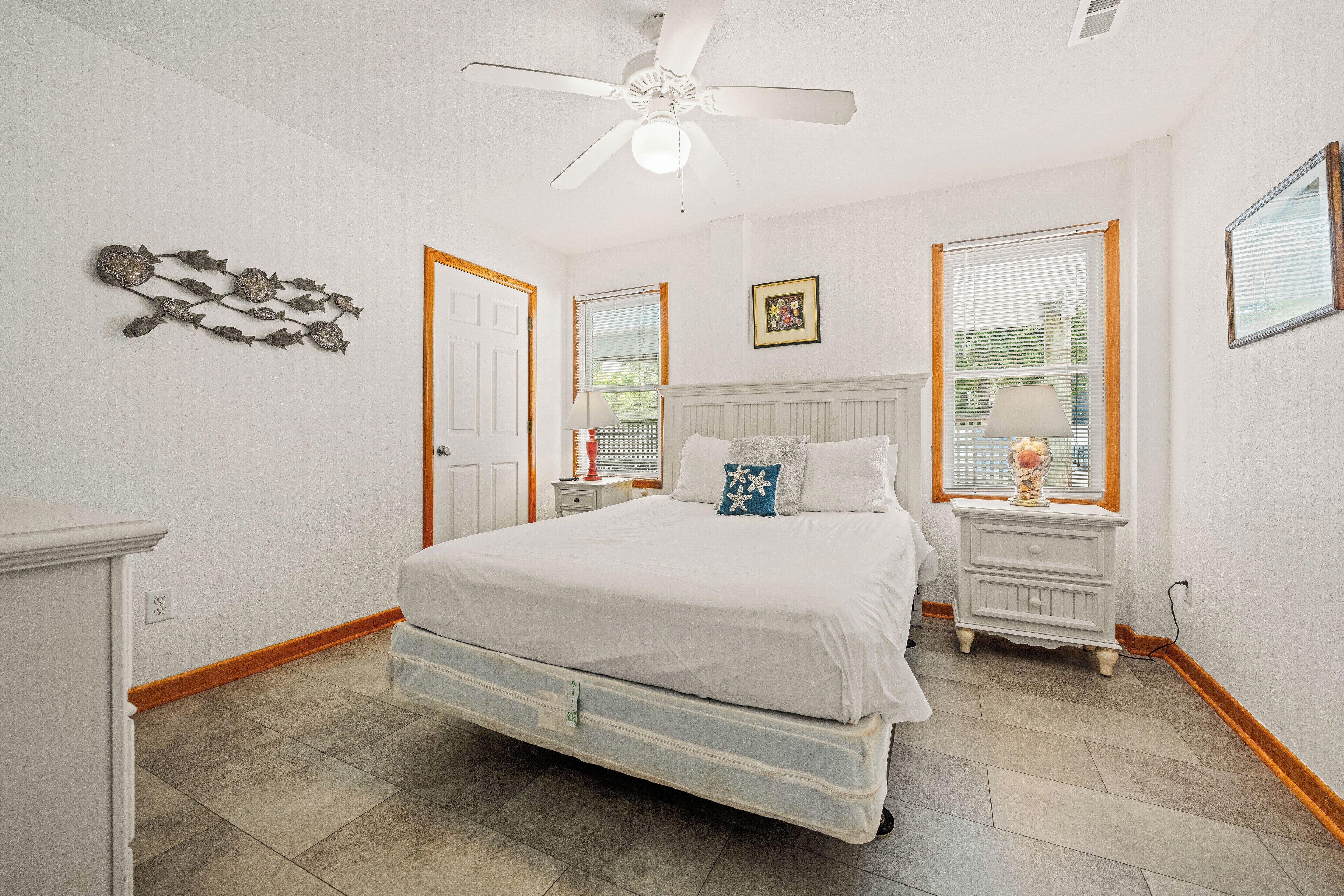 Une chambre à coucher comprenant un lit, une table de chevet, une lampe, un ventilateur de plafond et une fenêtre avec des stores.