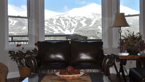 Un salón con un sofá de cuero, una mesa de centro con un cuenco decorativo, una lámpara y vistas a una montaña nevada a través de amplios ventanales.