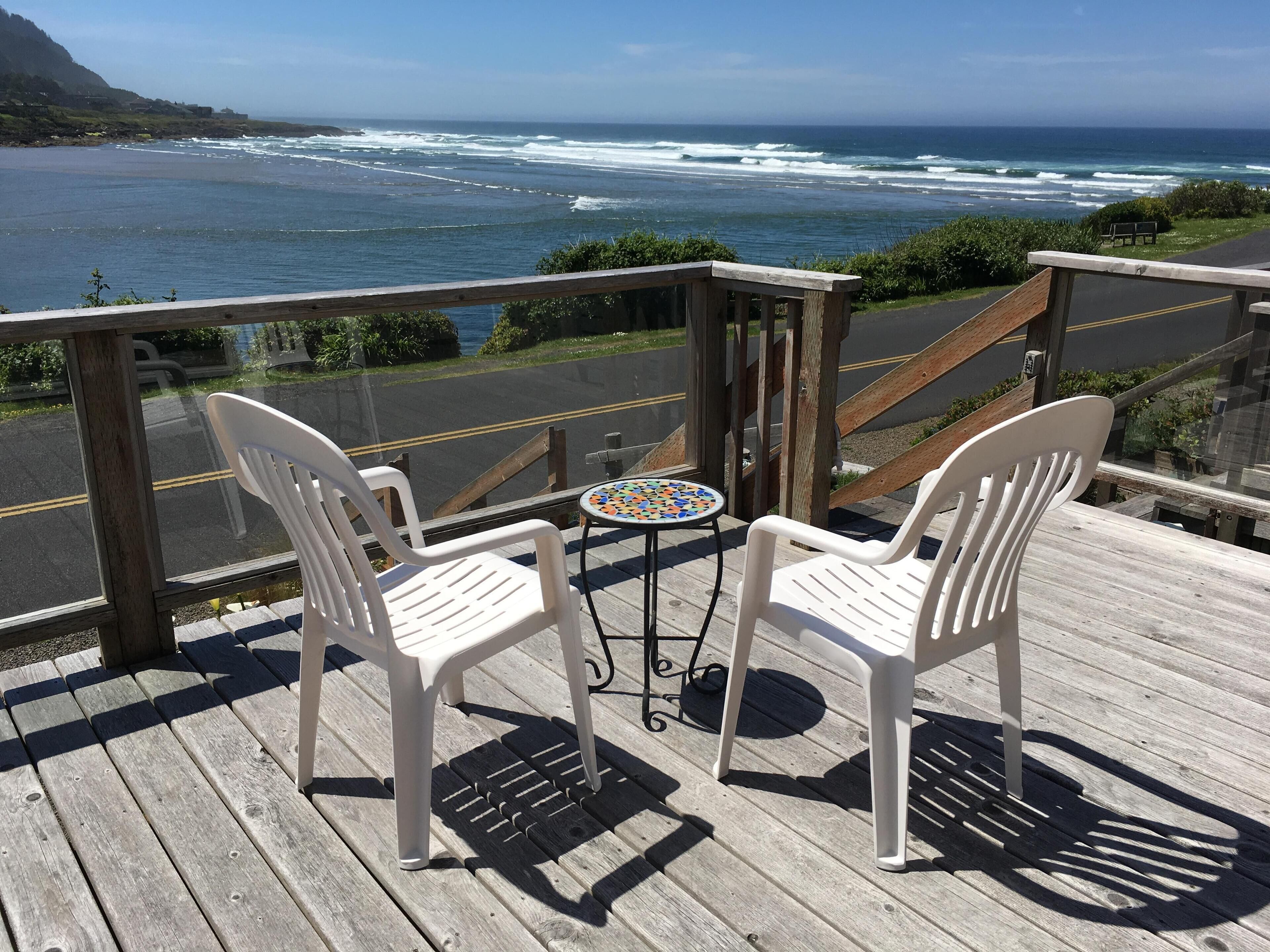 Une terrasse en bois avec deux chaises en plastique blanches et une petite table surmontée d’une mosaïque colorée, donnant sur une plage avec des vagues et une montagne lointaine.