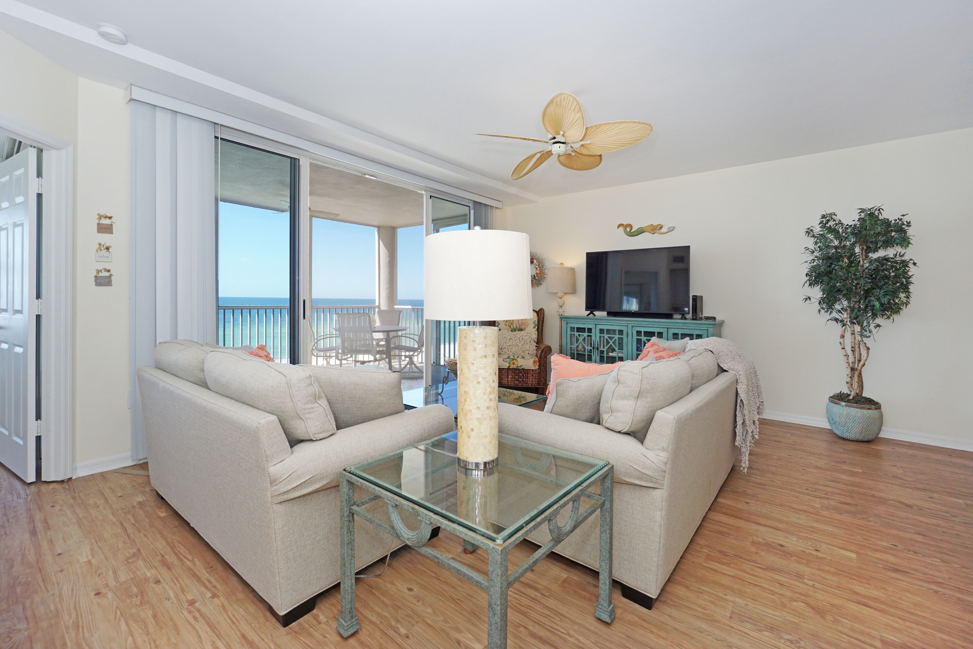 A living room with a glass coffee table, two sofas, a ceiling fan, a TV, a potted plant, and a view of the balcony.