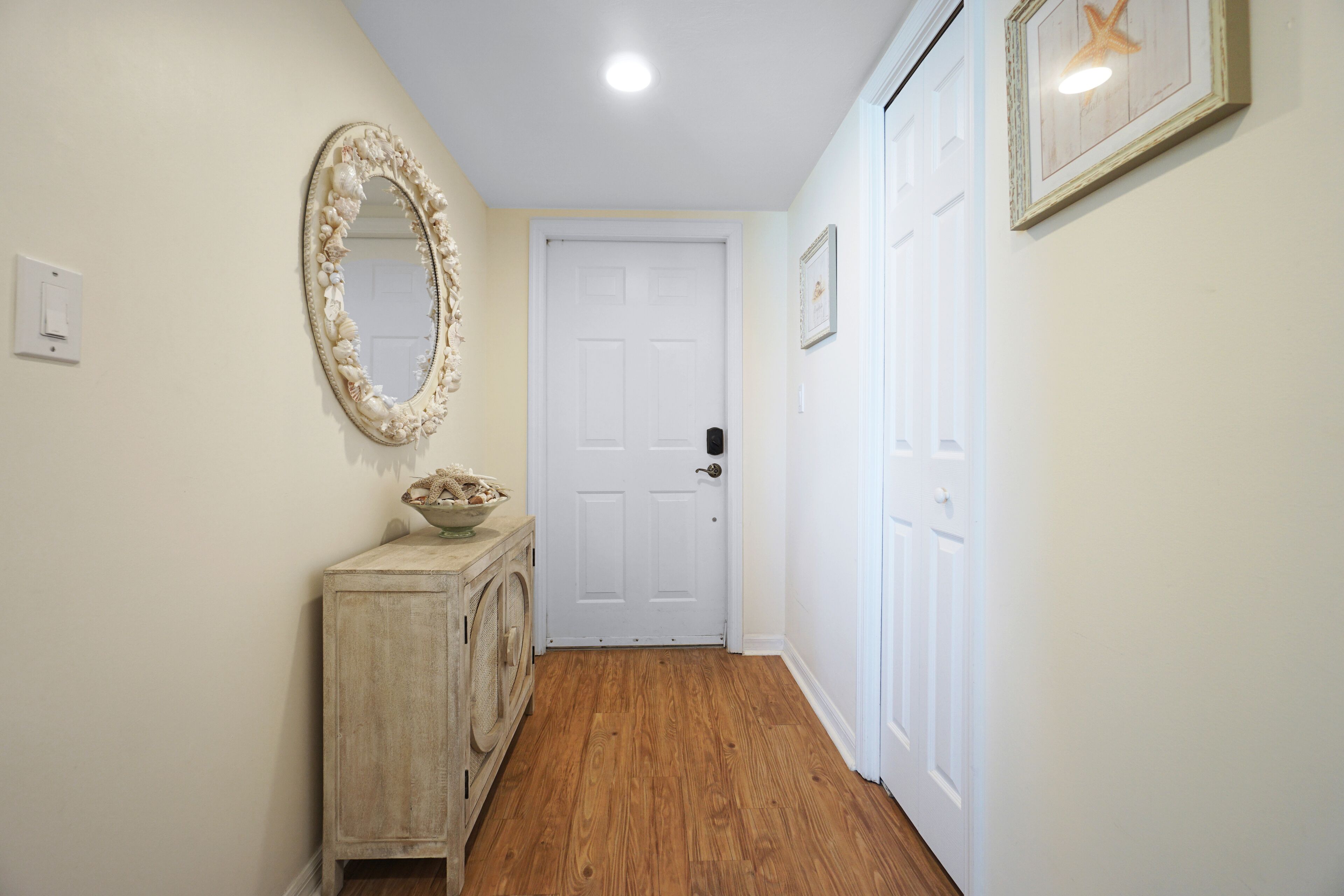 Un pasillo con suelo de madera, una puerta blanca, un espejo decorativo y un mueble pequeño con un cuenco encima.