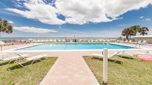 Piscina com espreguiçadeiras e palmeiras, cercada por um calçadão.
