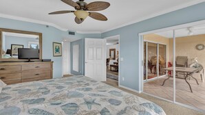 Une chambre à coucher comprenant un lit, un ventilateur de plafond, un téléviseur posé sur une commode et une porte-fenêtre coulissante donnant sur un coin repas.