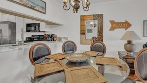 Une cuisine moderne avec des appareils en acier inoxydable, un coin repas doté d’une table en verre et de sets de table en fibres tressées, et un panneau décoratif représentant une plage accroché au mur.