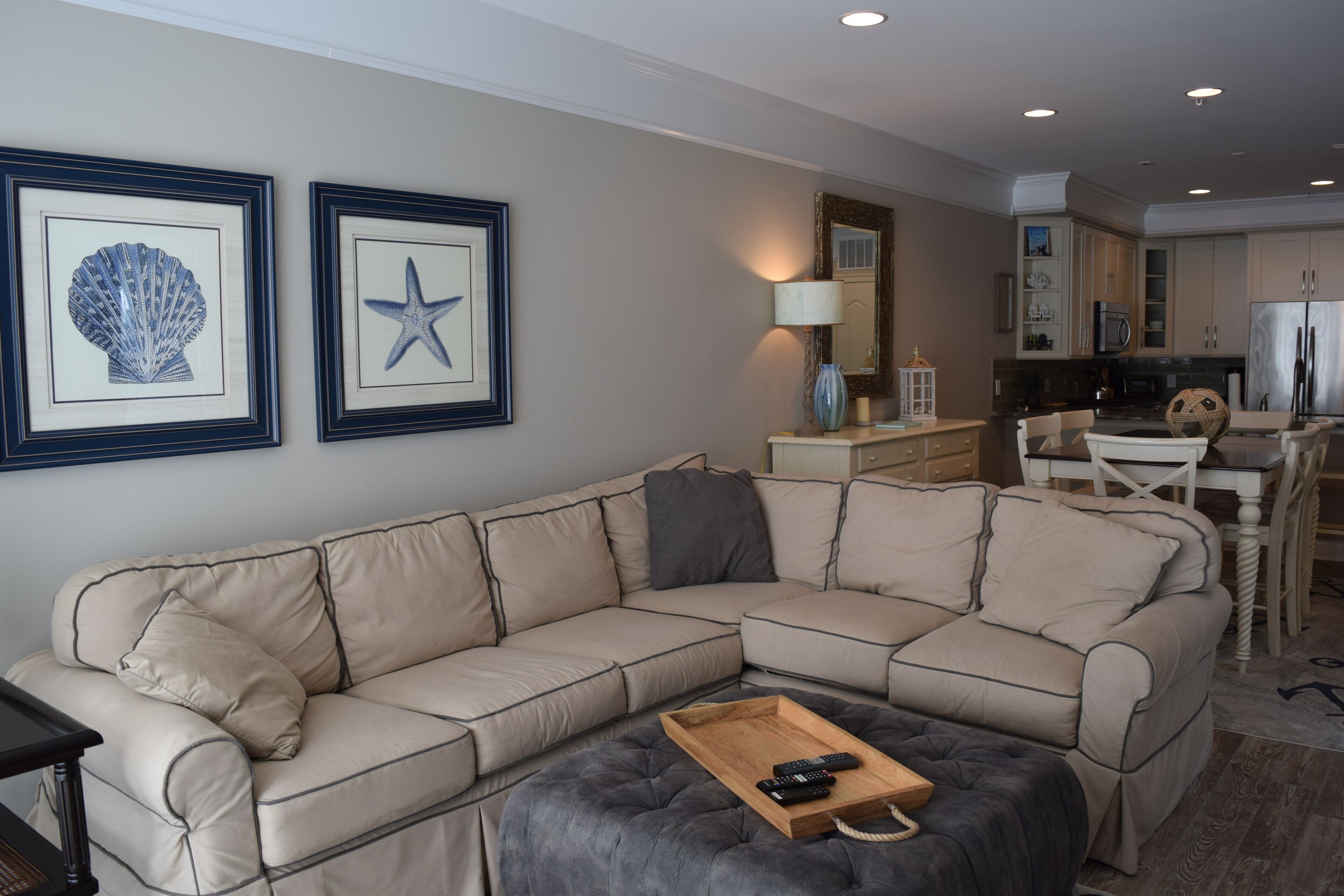 A beige sectional sofa with a wooden tray and remote controls, a dining area with a white table and chairs, and a kitchen with cabinets and appliances.