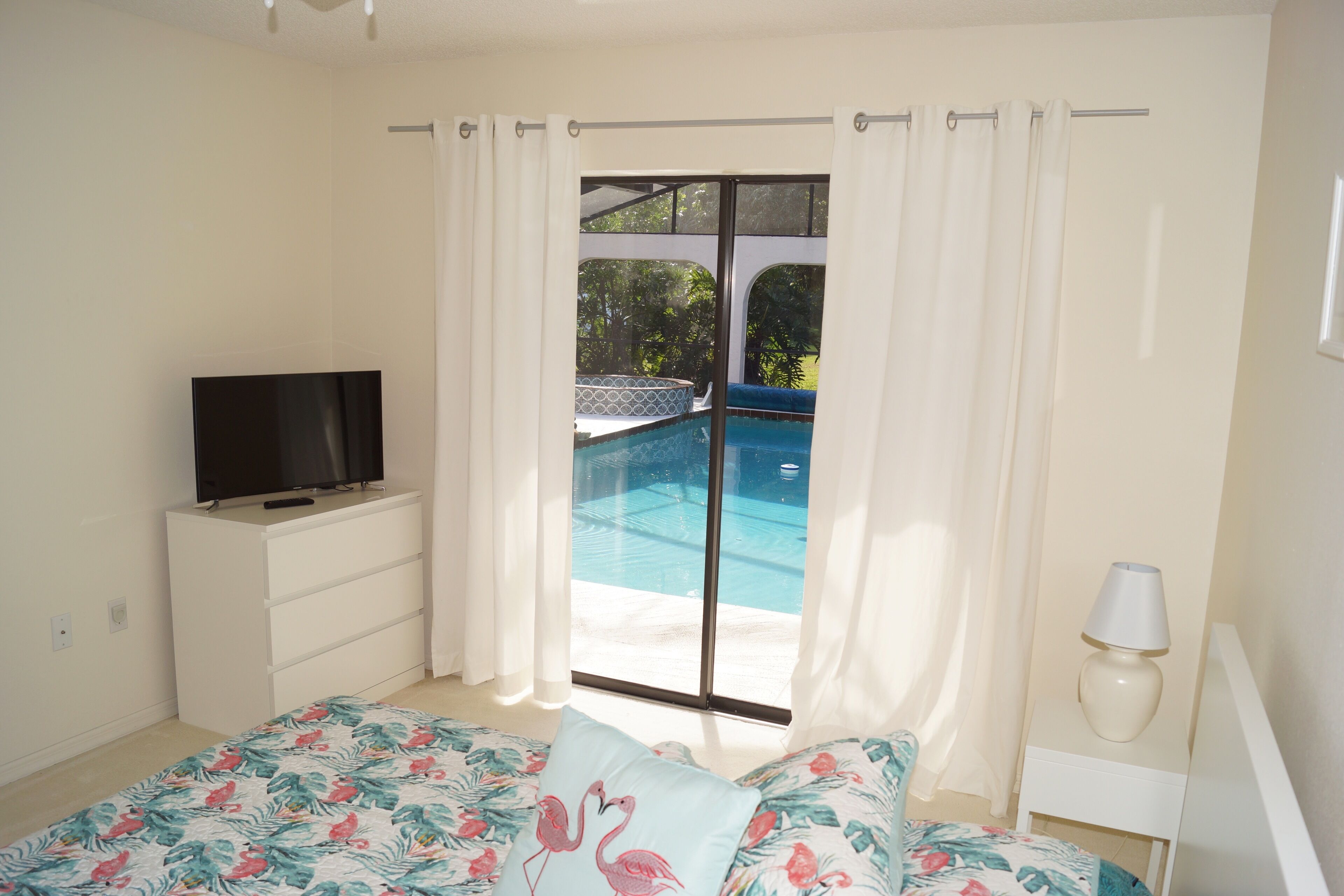 Ein Schlafzimmer mit einem Bett, einem Fernseher auf einer Kommode, einer Lampe und Blick auf einen Swimmingpool durch eine Glastür.