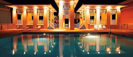Una piscina con reflejos, rodeada por un edificio de columnas y una zona cubierta con sillas.
