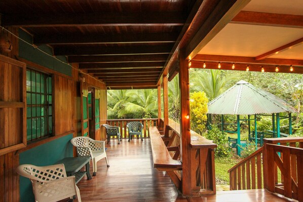 A wooden deck with outdoor seating, a covered area with a green umbrella, and a view of lush greenery.