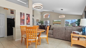 Uma sala de estar espaçosa com mesa e cadeiras de jantar, um sofá e uma mesa de centro. Há luminárias no teto, uma geladeira e uma janela com persianas.