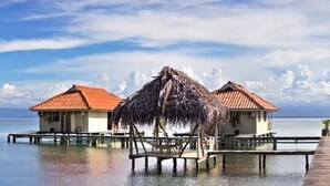 Eine Reihe von überwasserbungalows, verbunden durch eine Holzterrasse, jeweils mit einem Reetdach und Blick auf den Ozean.