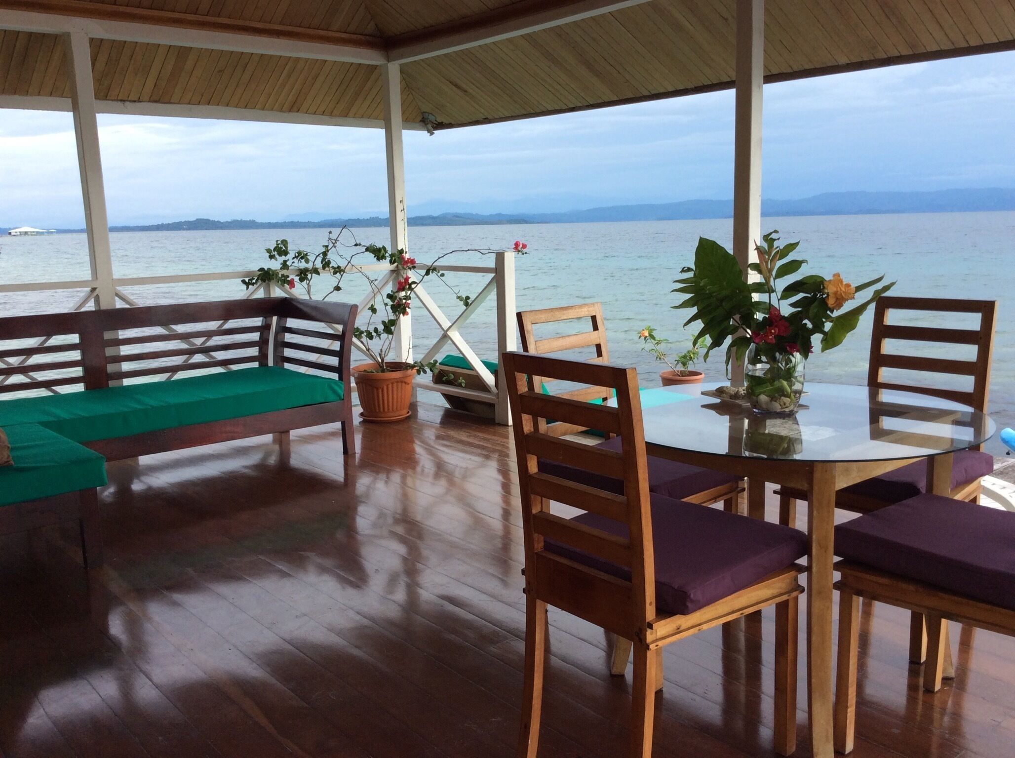 A covered outdoor seating area with wooden furniture and potted plants, overlooking a body of water.