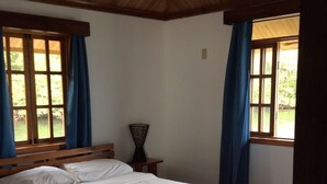 Ein Schlafzimmer mit Holzboden, einem großen Bett, einem Nachttisch und zwei mit Vorhängen versehenen Fenstern.
