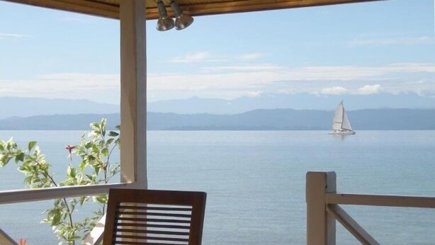 Eine Holzdielen-Terrasse mit Tisch und Stühlen, mit Blick auf ein ruhiges Meer, in der Ferne ein Segelboot und im Hintergrund Berge.