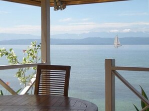Une terrasse en bois avec une table et des chaises, d’où l’on peut admirer une mer calme, avec un voilier à l’horizon et des montagnes en arrière-plan.