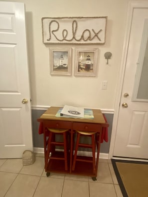 A hallway with a 'Relax' sign, two framed pictures of lighthouses, a small wooden table with papers, and a trash can.