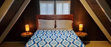 Un dormitorio con techo inclinado de madera, una cama con colcha estampada, dos lámparas de noche y una ventana con persianas.