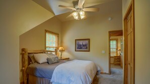 Une chambre avec un ventilateur de plafond installé sous une toiture inclinée, un lit avec une tête de lit, une table de chevet avec une lampe, un tableau encadré accroché au mur et une porte donnant sur une autre pièce.